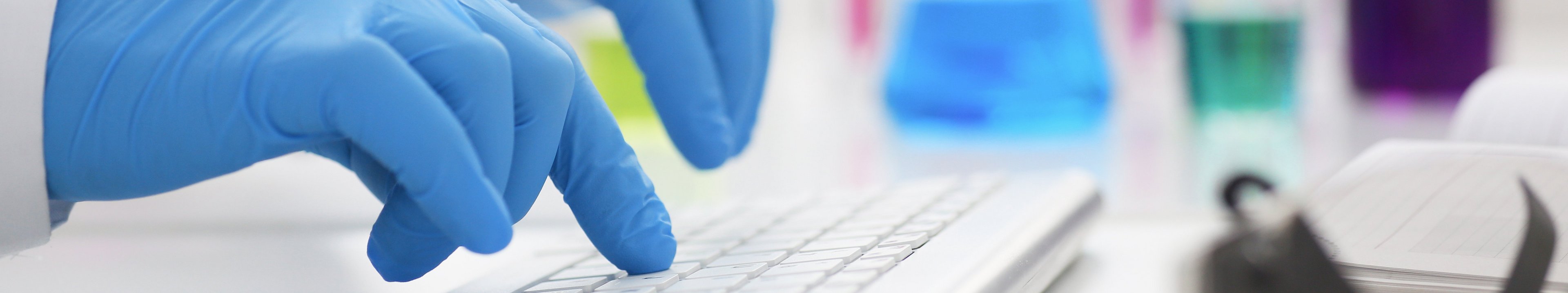 Hände in blauen Handschuhen tippen auf einer Tastatur, im Hintergrund farbige Reagenzgläser